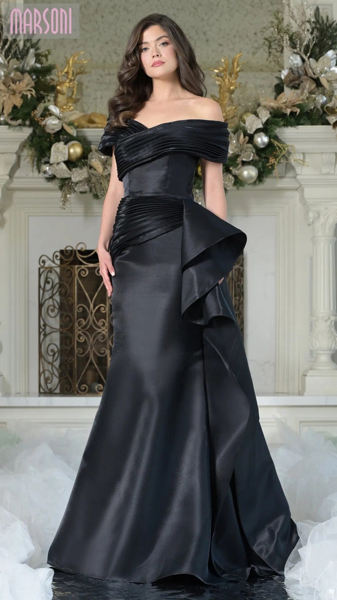 A woman in the Long Formal Dress: Marsoni MV1354 in black stands before a decorated fireplace adorned with floral and ornament arrangements.